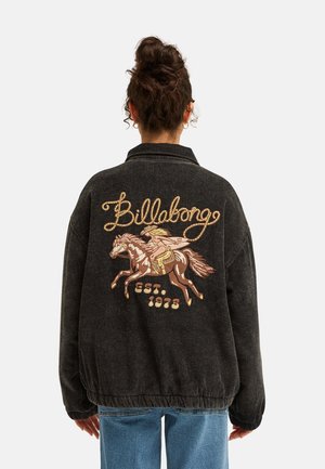 Person with hair in a bun wearing a black jacket with an embroidered horse and rider and the text "Billabong est. 1973" on the back.
