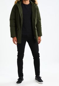 Grüne wattierte Parka-Jacke mit einem kapuzenbesetzten Pelzkragen, kombiniert mit einem schwarzen T-Shirt und schwarzen Slim-Fit-Hosen. Ausgestattet mit Reißverschluss und Knöpfen.