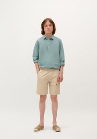 Polo shirt à manches longues bleu clair, shorts beiges avec poches et chaussures à enfiler couleur tan. Tissu doux, coupe décontractée et détails de conception subtils.
