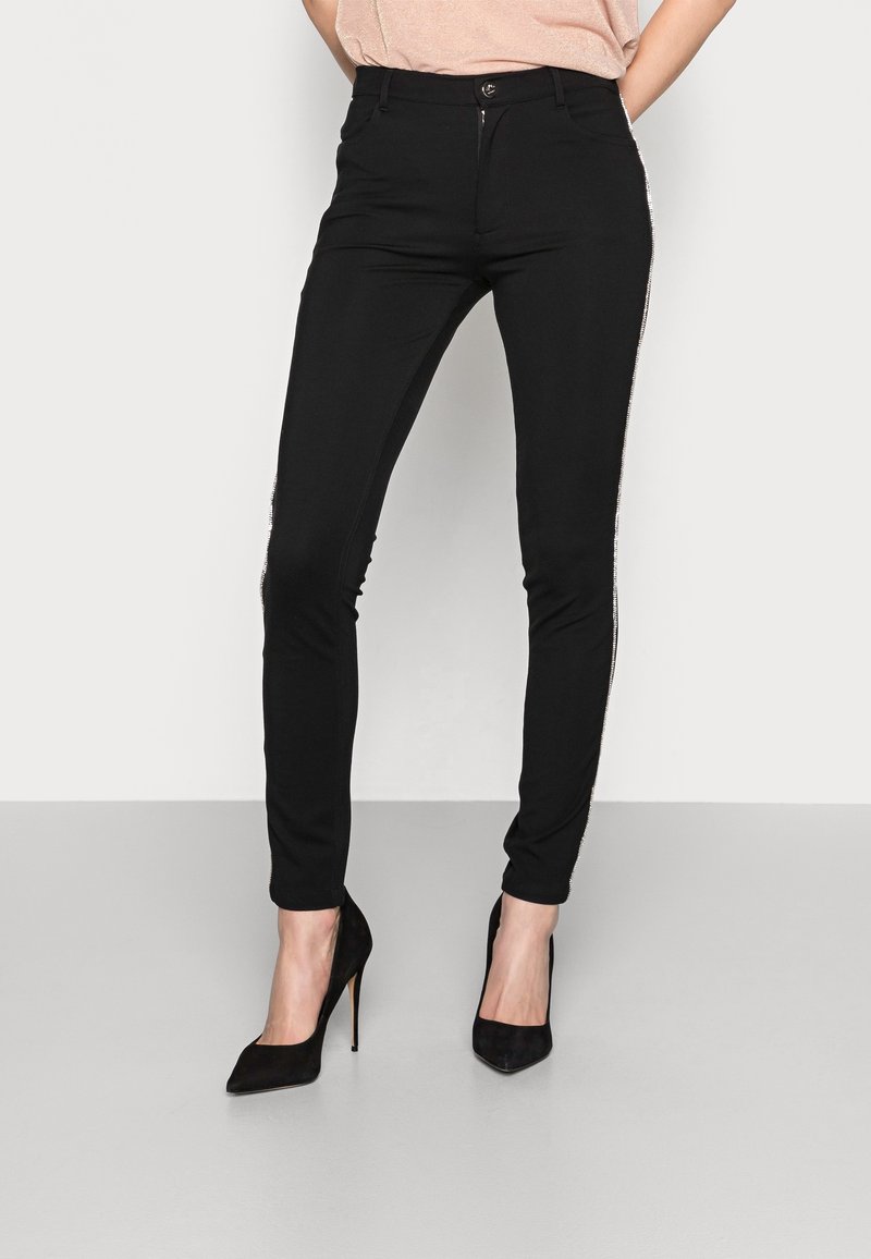 Svarta stretchbyxor med smal passform, med dragkedjedetalj längs de yttre sidorna. I kombination med svarta högklackade skor och en ljus topp.