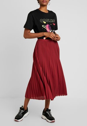Femme portant un t-shirt graphique noir "COSMIC", une jupe midi plissée rouge, des baskets noires et blanches, debout les mains jointes devant.