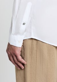 Weißes Hemd mit einem frischen Kragen und knöpfbaren Manschetten, das glattes Material zeigt. Beige strukturierte Hose, die unten sichtbar ist, mit subtilen vertikalen Linien.