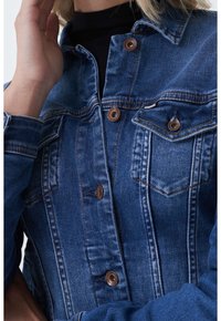 Denim jacket in dark blue with copper buttons, featuring a classic collar, chest pockets, and visible stitching details. Textured fabric.