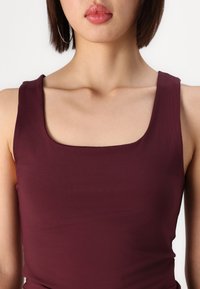 Haut sans manches bordeaux avec un col carré, fabriqué en tissu lisse, présentant une silhouette ajustée et un léger stretch.