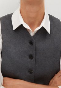 Szara wełniana kamizelka z pięcioma czarnymi guzikami, noszona na białej koszuli z kołnierzykiem. Kamizelka ma dopasowany krój i gładką fakturę.