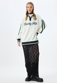 Felpa bianca con testo verde "Beverly Hills" e logo, abbinata a una lunga gonna nera di pizzo e stivali neri chunky.