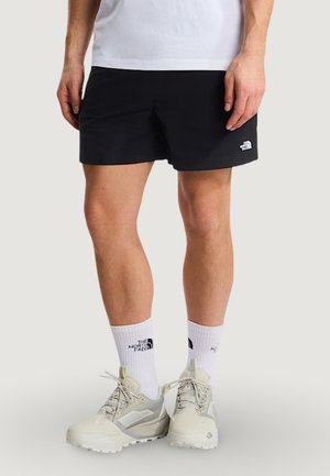 Uomo che indossa pantaloncini neri, calzini bianchi con marchio e scarpe da trail beige in piedi su uno sfondo neutro.