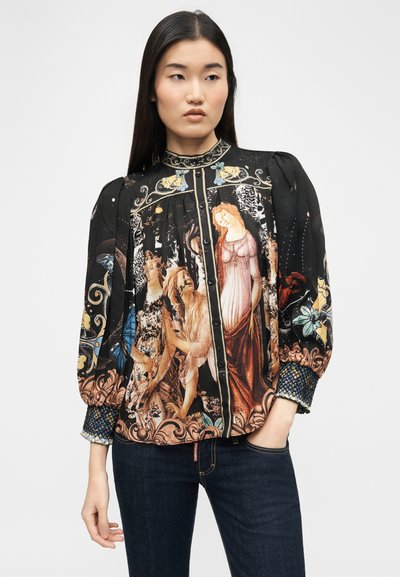 Femme portant un chemisier noir à boutons avec un imprimé d'œuvres d'art de style Renaissance et des poignets bleus à motifs, associé à un jean bleu foncé.