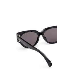 Ochelari de soare tip cat-eye negri cu lentile întunecate. Au rame din plastic elegante, cu accent de logo aurit și o finisare lucioasă. Brațele au o curbură netedă.