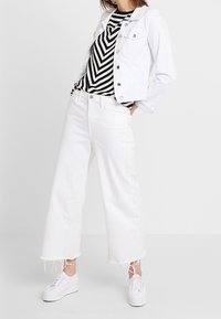 Bílá denimová bunda přes černobíle pruhovaný top, doplněná širokými bílými džíny s třepenými lemy a bílými teniskami.