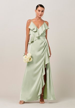 Kvinde holder hvide blomster iført lys grøn flæsetærklæd med slids, guldringe og sandaler mod en ensfarvet baggrund.