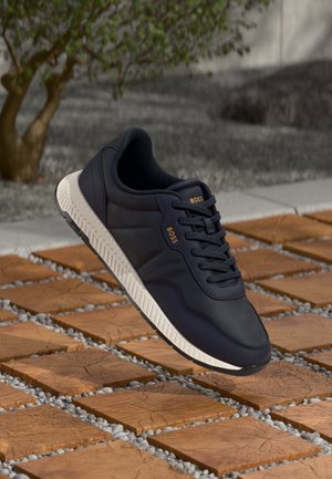 Czarny sneaker BOSS z białą podeszwą leżący na drewnianej płytce i ścieżce z kamyków na zewnątrz, w pobliżu małego drzewka.