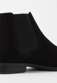 Bottine en daim noir avec des panneaux latéraux élastiques, un petit talon bloc et une finition lisse. Design simple avec des détails de couture minimalistes.