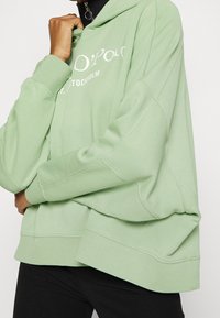 Sudadera verde de gran tamaño con una textura suave, que presenta un estampado gráfico en la parte delantera y una capucha con cordón. Pantalones negros visibles por debajo.