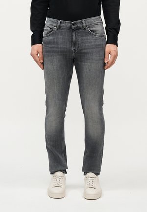 Άνδρας που φοράει ξεθωριασμένο γκρι τζιν κανονικής εφαρμογής, μαύρο μακρυμάνικο πουκάμισο και λευκά αθλητικά παπούτσια, στέκεται μπροστά σε ένα απλό φόντο.