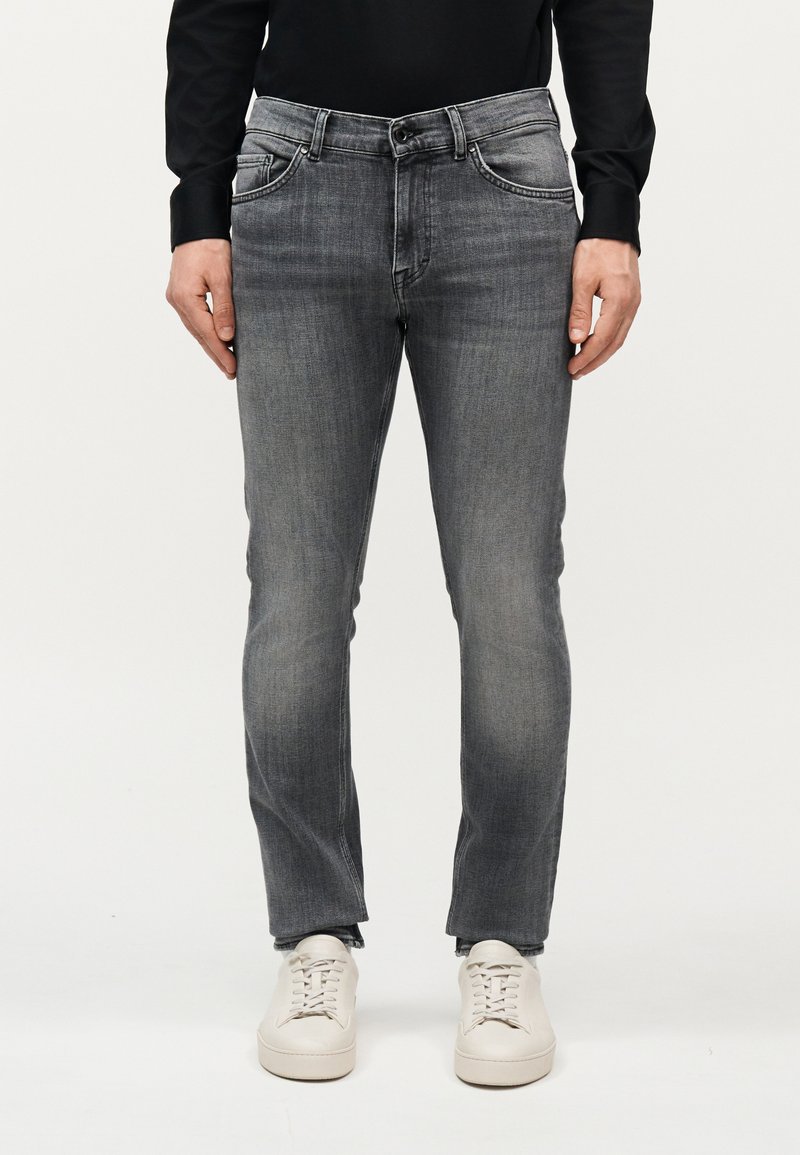 Άνδρας που φοράει ξεθωριασμένο γκρι τζιν κανονικής εφαρμογής, μαύρο μακρυμάνικο πουκάμισο και λευκά αθλητικά παπούτσια, στέκεται μπροστά σε ένα απλό φόντο.