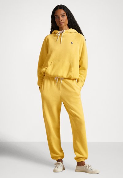Rumena jopa s kapuco in ujemajoče trenirke iz mehkega materiala, z žepom spredaj, z vrvico in rebrastimi manšetami. Nošeno z nevtralnimi supergami.
