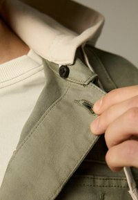 Roheline jakk heleda beeži krae ja musta nööbiga, millel on nähtav taskuava ja tekstuuriga kangas. Käsi hoiab kangast detaili.
