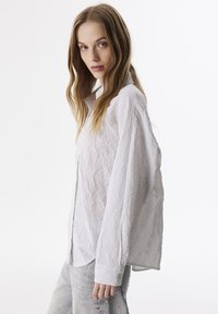 Camisa blanca de botonadura con textura y un sutil patrón a rayas. Presenta mangas largas y cuello de camisa. Llevada con pantalones de denim claros.