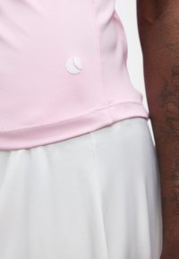 Κοντινό ενός ατόμου που φοράει ένα ανοιχτό ροζ πουκάμισο με λευκό λογότυπο και άσπρο παντελόνι, δείχνοντας το πλάι του κορμού και του μπράτσου.