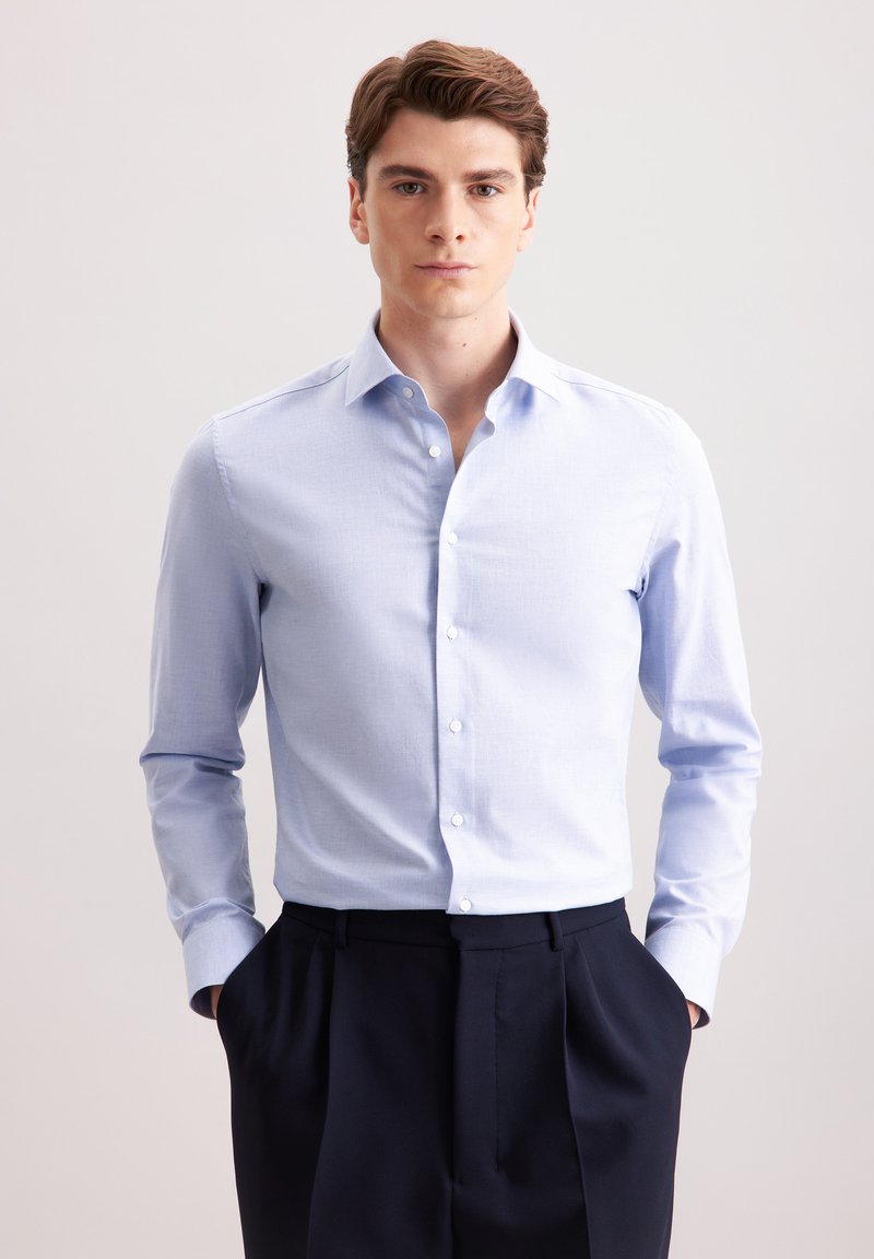 Camicia azzurra chiara con maniche lunghe in un tessuto morbido, abbinata a pantaloni blu navy. Collo classico e bottoni frontali.