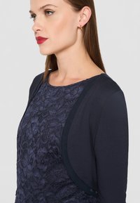Námořnický top s texturovanou krajkovou vrstvou, kulatým výstřihem a kontrastními hladkými rukávy. Zlaté kruhové náušnice doplňují design.