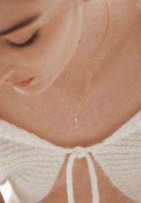Collar de oro con un colgante en forma de gota engastado con diamantes. La modelo lleva un acogedor suéter de punto blanco con un lazo en el escote.