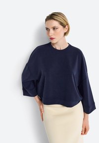 Felpa over-size blu navy con spalle cadenti, scollatura tonda e orlo a costine. Abbinata a una gonna aderente di colore chiaro. Texture liscia.