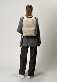 Beige ryggsäck med dubbla axelremmar, framficka med dragkedja och sidofickor. Materialet verkar strukturerat och har ett övre bärhandtag.