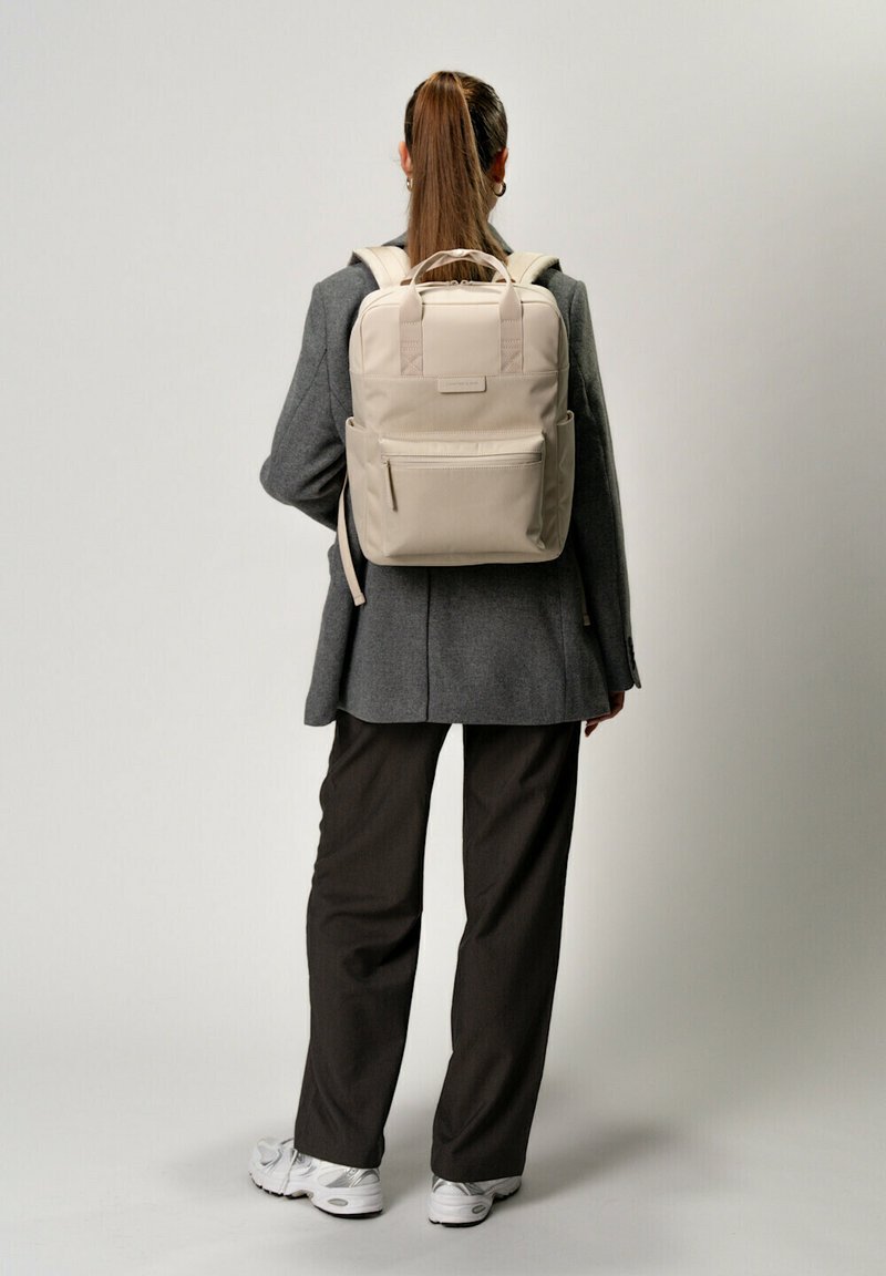 Beige ryggsäck med dubbla axelremmar, framficka med dragkedja och sidofickor. Materialet verkar strukturerat och har ett övre bärhandtag.