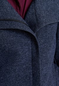 Cappotto blu scuro con texture, caratterizzato da un colletto prominente e una cerniera frontale. L'interno è foderato di bordeaux, mostrando un design a strati.