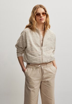 Mujer con chaqueta beige texturizada con cremallera, pantalones beige a juego, gafas de sol y collares de plata superpuestos, de pie con las manos en los bolsillos.