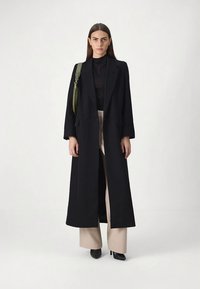 Sieviete, tērpusies garā melnā mētelī virs melnas blūzes un bēšiem platām biksēm, nēsā zaļu plecu somu un stāv pretī vienkāršai baltai fona.