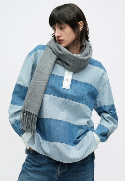 Chemise en denim rayée bleue avec une patte de boutonnage blanche, manches longues et une poche latérale, accompagnée d'une écharpe grise à franges.
