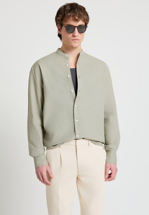 Junger Mann mit runden schwarzen Sonnenbrillen, hellgrauem Hemd mit Mandarin-Kragen und cremefarbenen Hosen, stehend vor weißem Hintergrund.