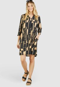 Schwarz-beiges, gemustertes Kleid mit Relaxed Fit, Dreiviertelärmeln, Kragen und seitlichen Taschen. Getragen mit schwarzen Sandalen.