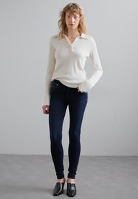 Witte geribde langetermijn top met een kraag, gecombineerd met donker skinny jeans en zwarte instap schoenen. De outfit is minimalistisch en aansluitend.
