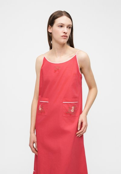 Γυναίκα που φοράει κόκκινο φόρεμα χωρίς μανίκια με λεπτές τιράντες και κεντήματα με λουλούδια στις τσέπες και το στήθος, στέκεται μπροστά σε λευκό φόντο.