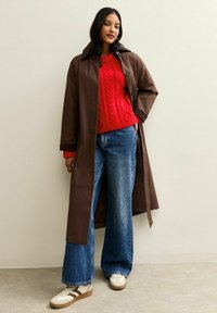 Bruine waterdichte trenchcoat over een rode kabelgebreide trui, gecombineerd met wijde blauwe denim jeans en witte sneakers met bruine accenten.