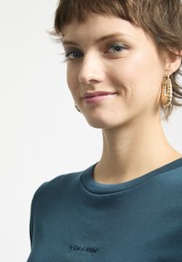 Tee aus türkisfarbenem Baumwollmaterial mit rundem Halsausschnitt und kurzen Ärmeln, ausgestattet mit einem kleinen gestickten Logo in Brusthöhe. Glatte Textur.