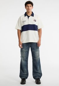 White polo shirt with a navy horizontal stripe, short sleeves, and embroidered details. Paired with loose blue cargo pants featuring side pockets.