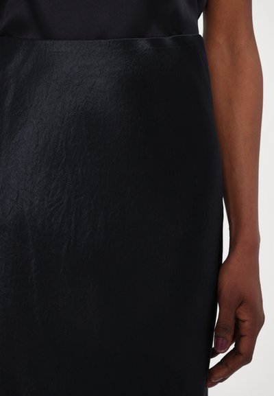 Črna krila z nagubano teksturo, tesno prileganje in gladkim pasom. Posnetek od blizu, kjer je roka naslonjena na stran, z nežno barvo nohtov.