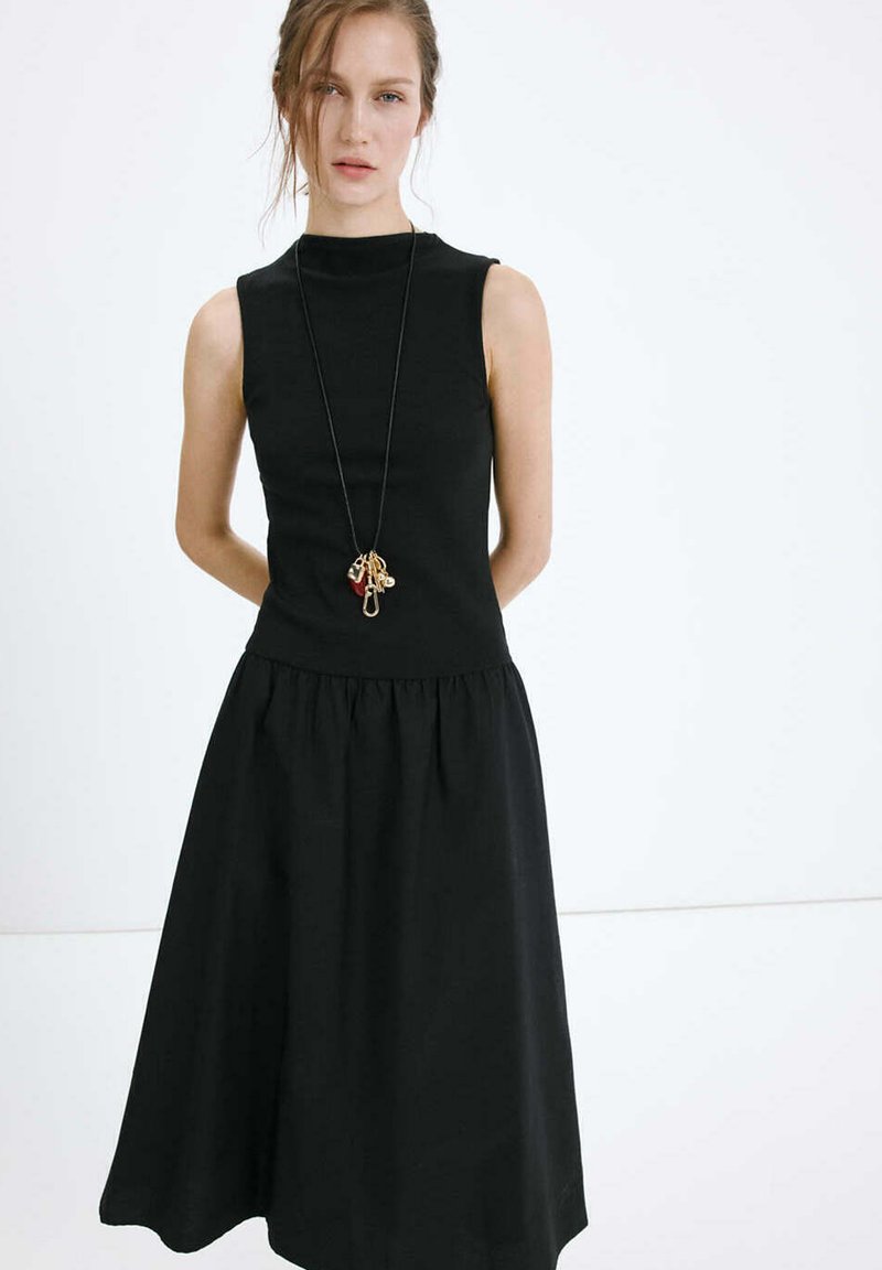 Jeune femme debout sur fond blanc, portant une robe noire sans manches et un long collier avec des breloques dorées et rouges.