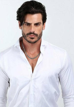 Camicia bianca con bottoni e colletto, design aderente. Collana grigia poggia su un petto nudo, capelli scuri e mossi, e barba curata sono visibili.