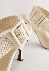 Sandales à talons couleur crème avec un design de lanières tissées, une boucle carrée en métal, et un talon fin avec une base noire contrastante.