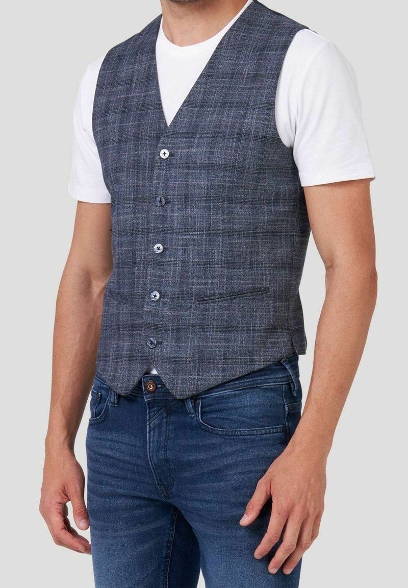 Homme portant un gilet à carreaux gris boutonné sur une chemise blanche à manches courtes et un jean bleu sur un fond uni.