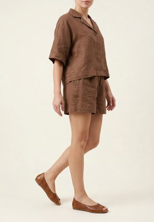 Frau trägt braunes kurzärmeliges Hemd mit Kragen und passende Shorts mit perforierten braunen flachen Schuhen, steht vor hellem Hintergrund.