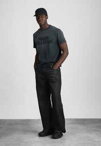 Mann mit schwarzer Mütze, verblasstem schwarzen T-Shirt mit dem Text "PSEUDO INTELLECTUAL", lockeren schwarzen Jeans und schwarzen Turnschuhen, der mit den Händen in den Taschen steht.