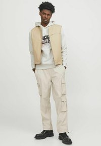 Beige ærmeløs vest, grå hættetrøje med grafisk tryk, beige cargo-bukser med flere lommer, sorte støvler, teksturerede stoffer.