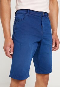 Siniset puuvillashortsit, joissa on muotoiltu vyötärö, suorat lahkeet ja käärityt hihnat. Sisältää etutaskut ja yhden napin kiinnityksen.